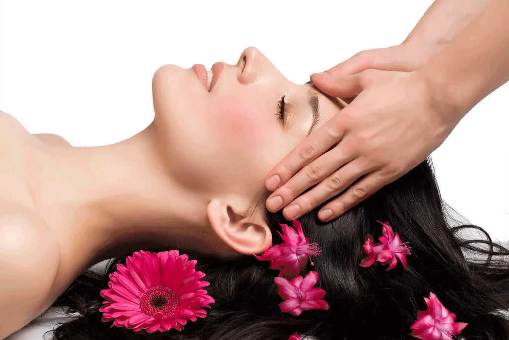 Une femme aux longs cheveux noirs allongée avec des gerberas roses pour un massage crânien et visage de 25 minutes