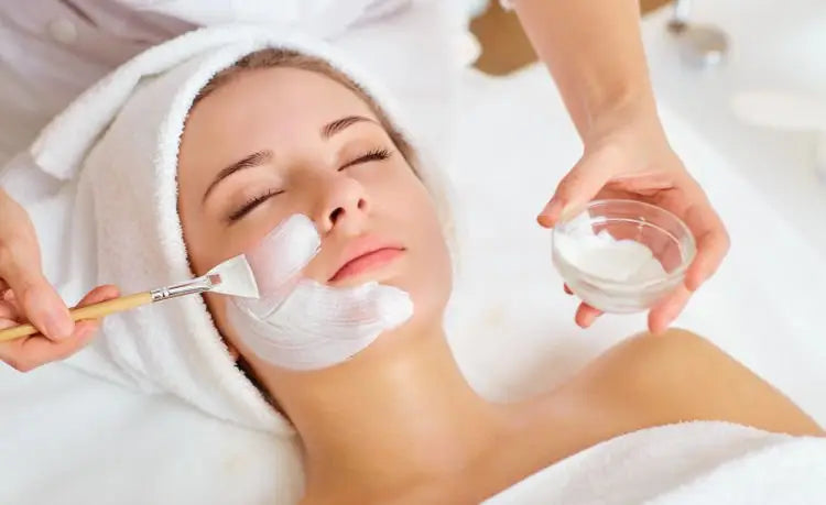 Une femme aux yeux fermés reçoit un soin visage purifiant avec un masque blanc appliqué au pinceau lors du nettoyage de peau